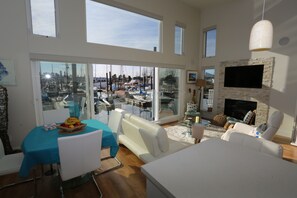 A spacious living area with a large glass door leading to a marina view, a flat-screen TV mounted on a stone wall, and a dining area with a blue tablecloth.