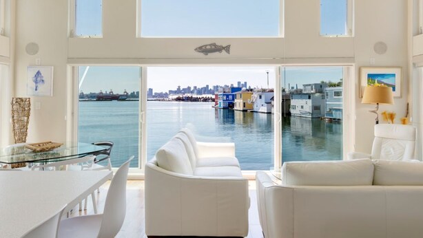 Un intérieur moderne avec une table à manger en verre, des canapés blancs et une grande fenêtre donnant sur une marina avec des bateaux et une vue sur les gratte-ciel de la ville.