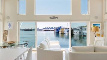 Un intérieur moderne avec une table à manger en verre, des canapés blancs et une grande fenêtre donnant sur une marina avec des bateaux et une vue sur les gratte-ciel de la ville.