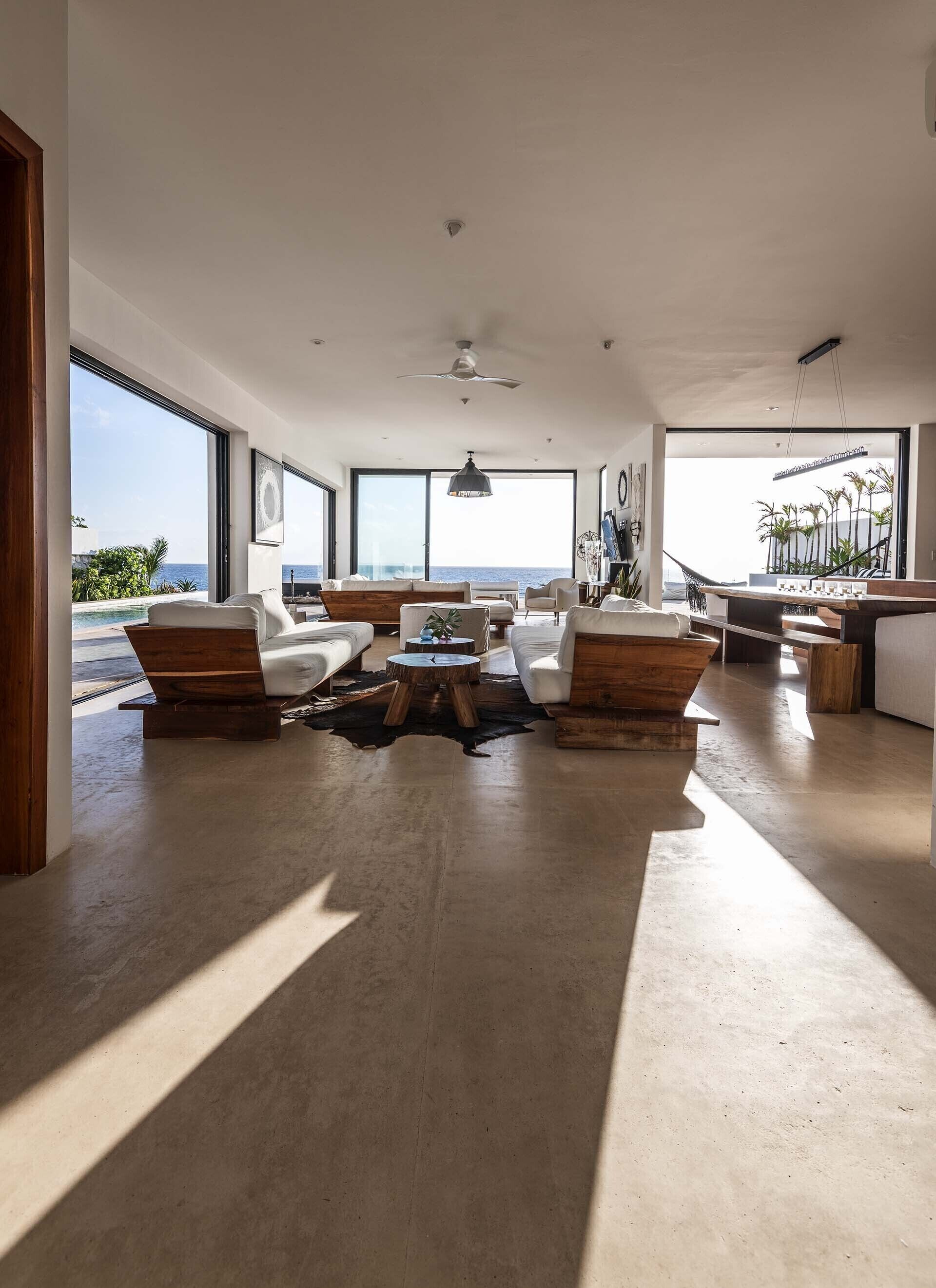 Un ampio soggiorno con una grande finestra vista piscina e sull'oceano, arredato con mobili moderni e un tavolino da caffè in legno al centro.