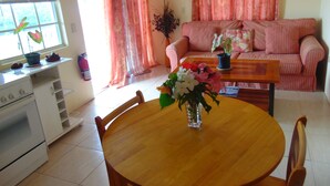 A living room with a sofa, a dining table with chairs, a kitchen area with a stove, and a window with curtains. - Antigua Seaview Alamanda 6 - Affordable, Convenient And Comfortable (St. John's)