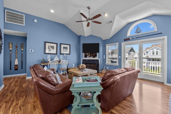 A living room with a fireplace, leather sofas, a ceiling fan, and a view of houses through large windows.