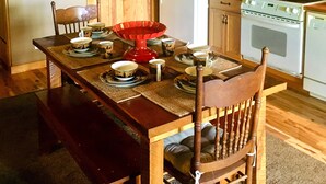 Une table à manger en bois, surmontée d’un lustre, est entourée de chaises et agrémentée d’un centre de table rouge. On aperçoit une horloge et des meubles en bois à l’arrière-plan.