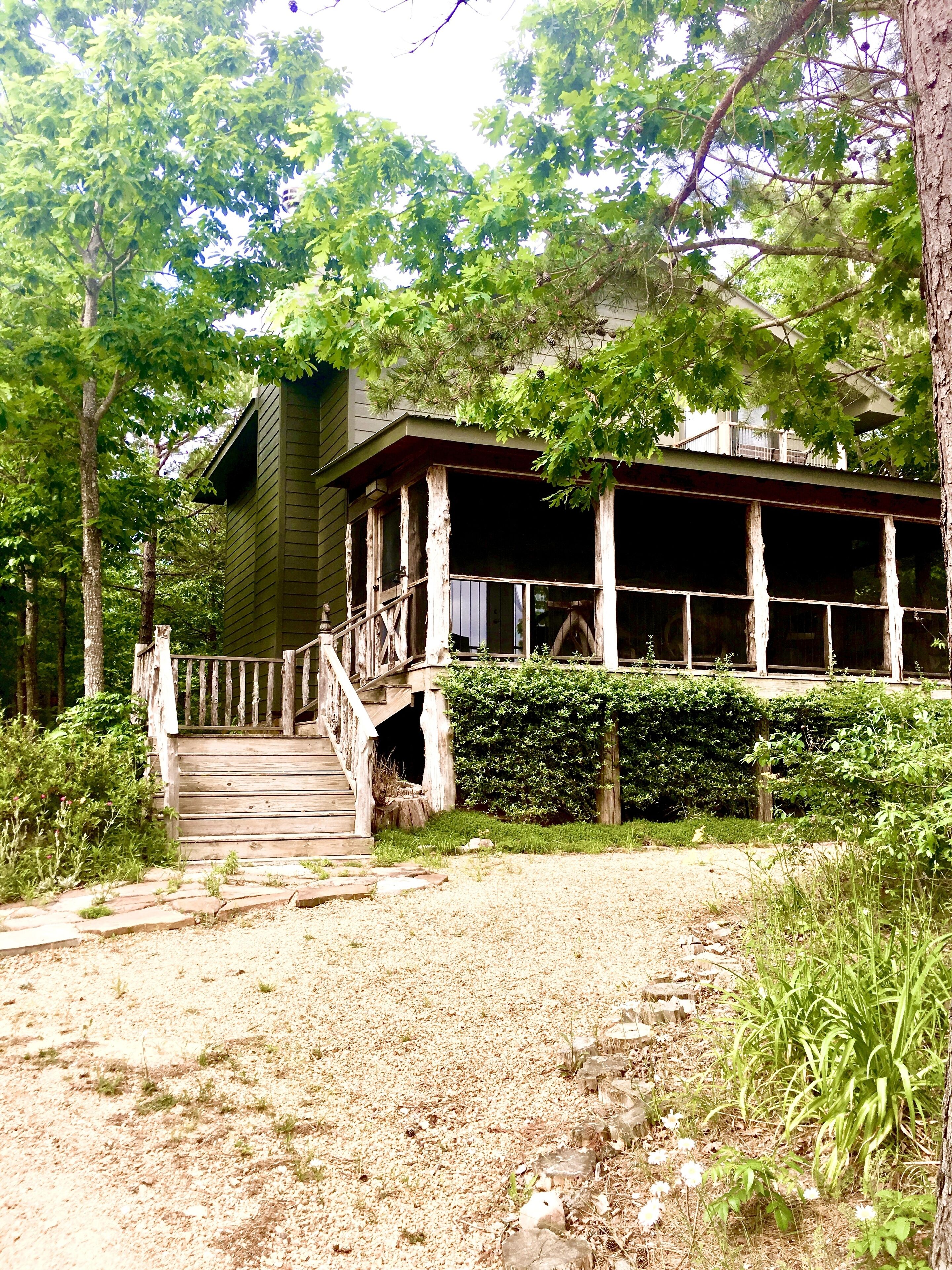 Une maison en bois à l’allure rustique, dotée d’un porche et entourée d’une végétation luxuriante et d’arbres.