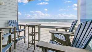 Eine Holzterrasse mit Adirondack-Stühlen und einem kleinen runden Tisch mit Blick auf einen Strand und einen klaren Himmel.