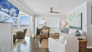Ein geräumiges Wohnzimmer mit einem weißen Sofa, einem Korbstuhl und einem Couchtisch. Ein Deckenventilator ist sichtbar, und ein großes Fenster bietet einen Blick ins Freie.