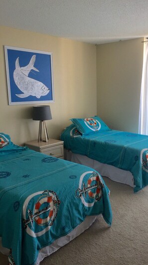 Quarto com duas camas, uma mesa de cabeceira, abajur e um quadro na parede. O quarto possui uma varanda acessível de todos os cômodos.