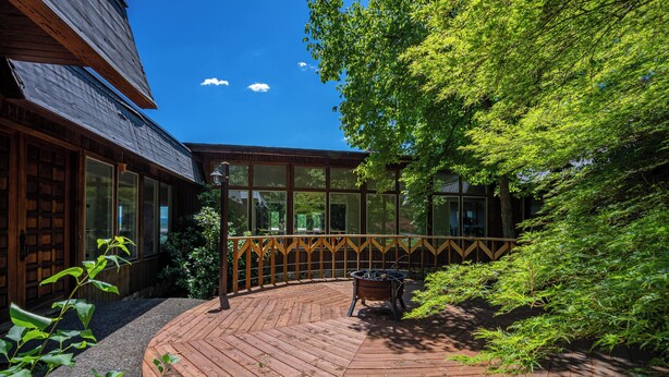 Une terrasse en bois avec un foyer, entourée d’une végétation luxuriante et d’un bâtiment en bois doté de grandes fenêtres.