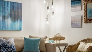 En stue med sofa, sofabord og fletstol. Der er hængende lamper, et maleri og et indrammet billede på væggen.