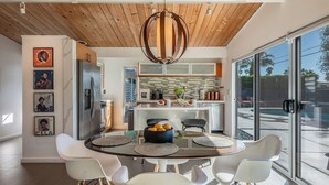Una cocina moderna con techo de madera, un comedor con cristales y sillas blancas, y vista a una piscina a través de grandes ventanales.