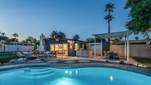 Un área de piscina moderna al aire libre con un espacio de estar cubierto, un patio y vistas a una casa con garaje.
