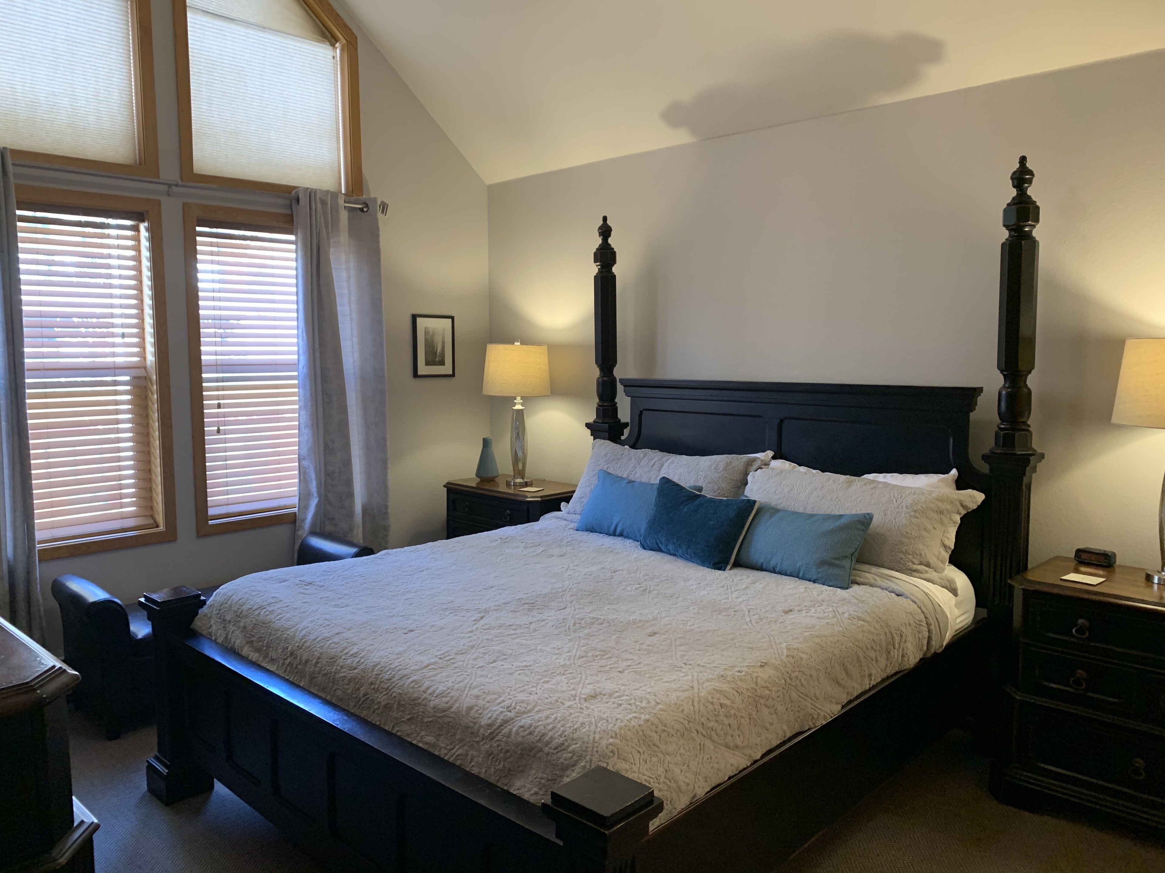 A neatly made bed with a headboard, two bedside tables with lamps, a window with blinds, and a small picture on the wall.