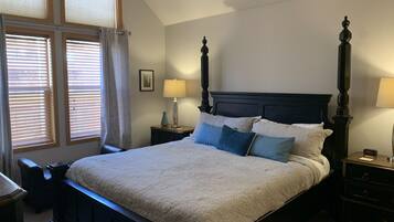 A neatly made bed with a headboard, two bedside tables with lamps, a window with blinds, and a small picture on the wall.