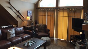 Una sala de estar acogedora con un sofá de cuero marrón, una mesa de centro con adornos, un televisor y un ventanal con cortinas.