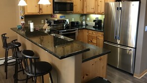 Cocina con armarios de madera, refrigerador de acero inoxidable, microondas, cafetera y un lavamanos. Hay tres taburetes negros.