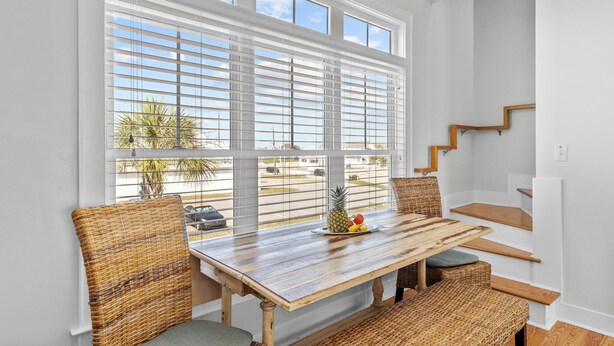 A dining area with a wooden table and wicker chairs, a window with blinds, and a staircase. - Poolside House w/ Ocean Views, Steps from Beach & Restaurants (Atlantic Beach)
