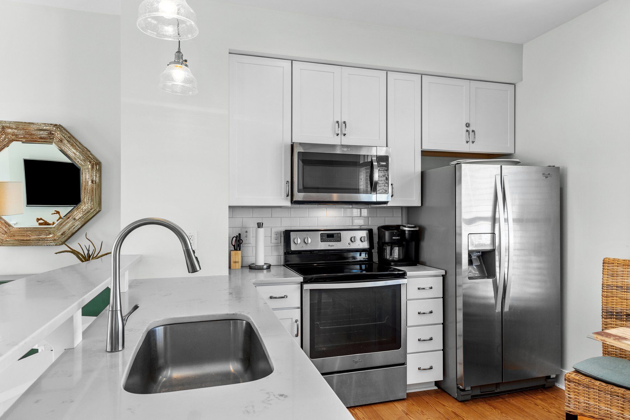 A modern kitchen with white cabinets, stainless steel appliances, a sink, and a dining area with a wicker chair.