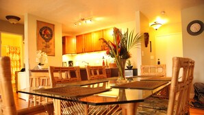 A dining area with a glass-top table surrounded by wicker chairs, a kitchen in the background, and a clock on the wall. - Shores of Maui - Ocean View- Top Floor Unit--Spacious! AC & WiFi (Kihei)