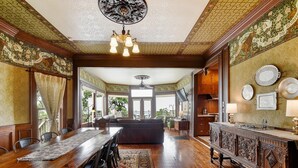 A dining room with a long wooden table, chairs, and a chandelier. There is a sofa and a television in the background. - Charming Victorian Home w/Pickle Ball Court, Pool, Hot Tub, Game Room (San Marcos)