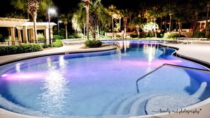 A nighttime swimming pool with a curved edge, surrounded by palm trees and a covered seating area.