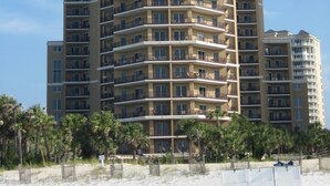 A multi-story hotel with balconies, located near a sandy beach with palm trees.