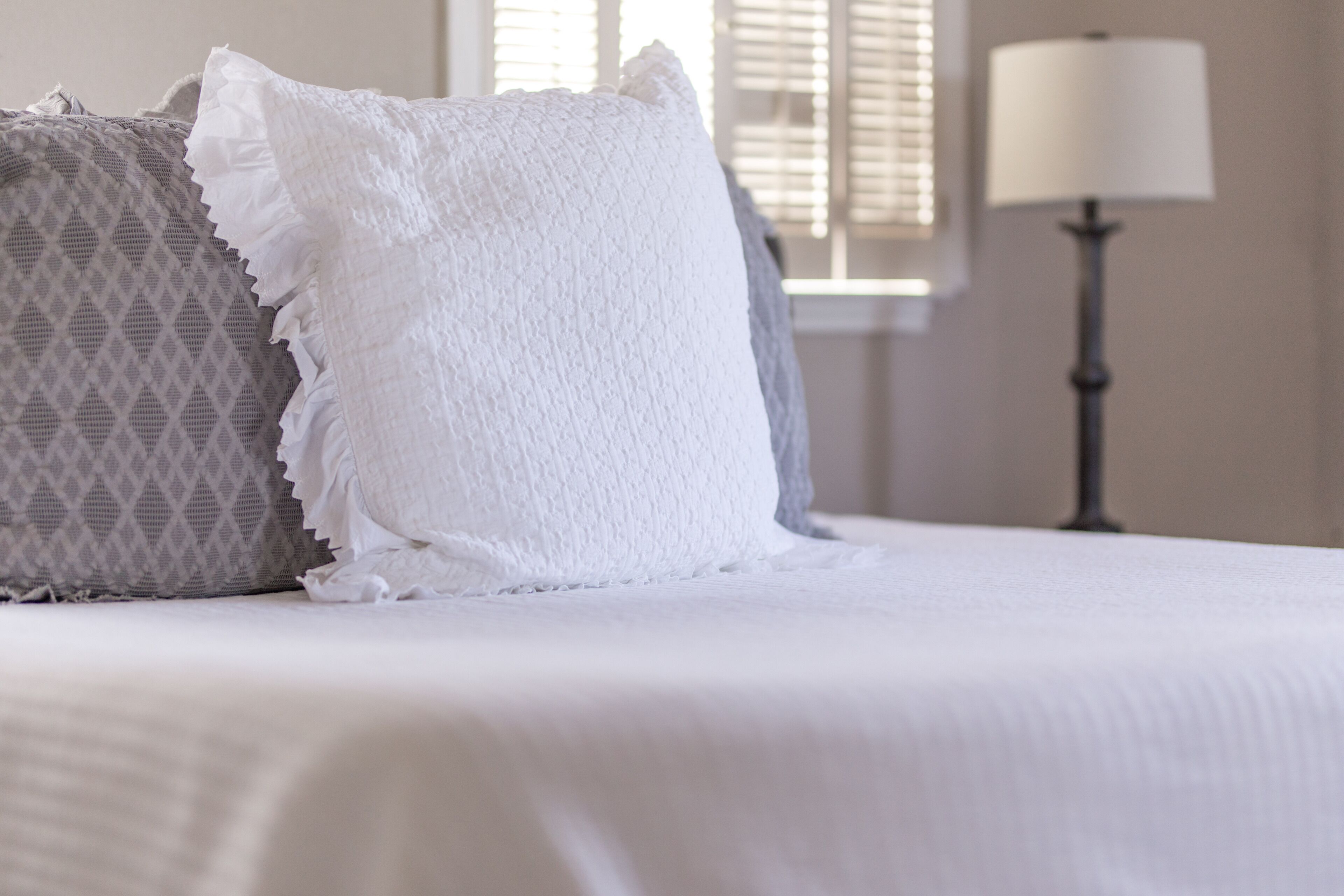Una cama con almohadas blancas y estampadas, una lámpara y una ventana con persianas al fondo.