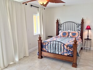 A bedroom with a wooden bed frame, patterned bedding, a window with curtains, a red lamp, and a ceiling fan.