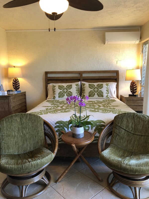 A bedroom with a bed, two green upholstered chairs, a small table with a potted plant, a ceiling fan, and wall-mounted lamps.
