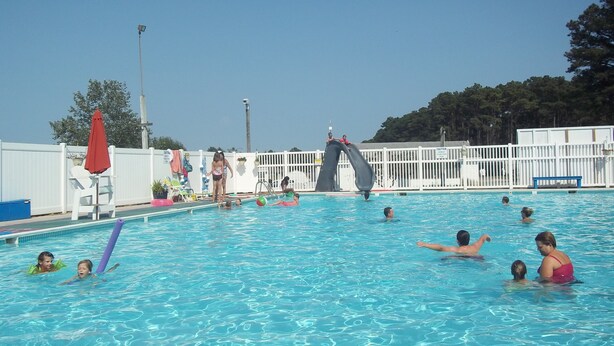 A swimming pool with a slide, surrounded by a white fence, and trees in the background.