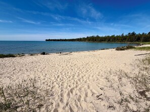 Ein sandiger Strand mit Fußabdrücken, einem klaren blauen Himmel und einer bewaldeten Küstenlinie.
