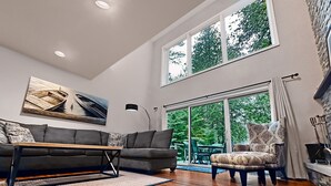 Ein geräumiges Wohnzimmer mit einem grauen Sofa, einem gemusterten Sessel und einem Holztisch. Ein großes Fenster bietet Blick auf viel Grün.