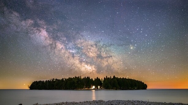 Ein sternenklarer Nachthimmel über einem stillen See mit einer bewaldeten Insel im Hintergrund.