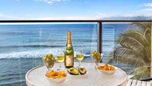 A table with a bottle of champagne, two glasses, bowls of grapes and chips, and a view of the ocean.