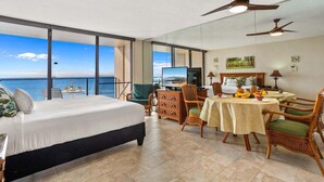 A hotel room with a large bed, a dining table with chairs, a TV, and a view of the ocean.