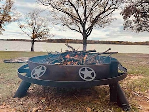 A metal fire pit with star designs, surrounded by grass and trees, near a body of water.