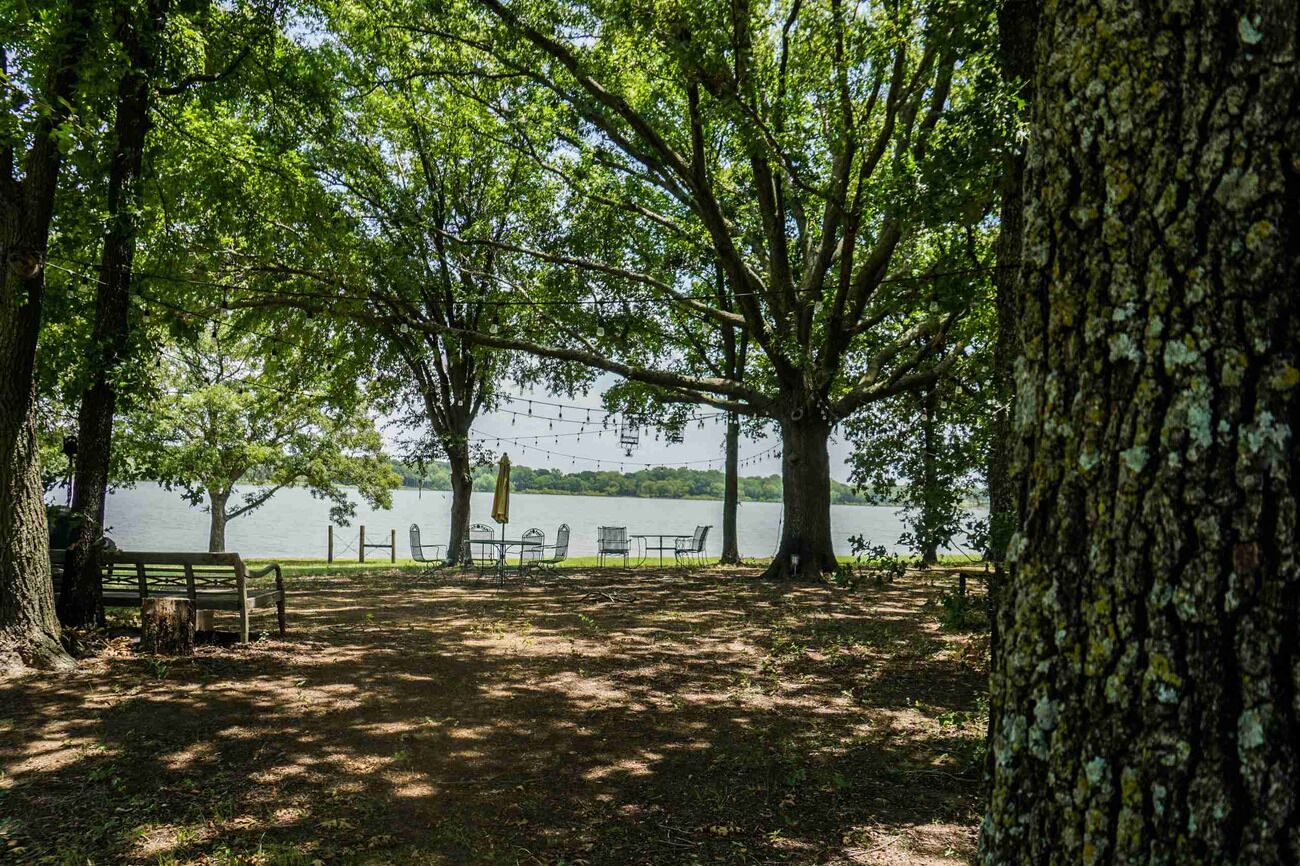 A park with a bench, tables, and chairs under trees by a lake.