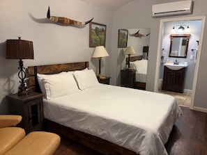 A hotel room with a large bed, bedside lamps, a wooden headboard, a nightstand, a small table, a mirror, a bathroom with a sink and mirror, and a wall-mounted air conditioner.