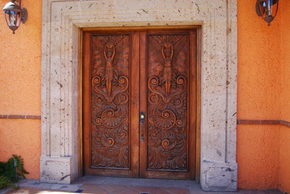 A double wooden door with intricate carvings, flanked by two wall-mounted lanterns, set in a stucco wall with a stone base.