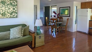 Una sala de estar con un sofá verde, una mesa de comedor con sillas, un ventilador de techo y una cocina con refrigerador.