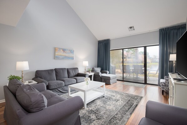 A modern living room with grey sofas, a white coffee table, and a large sliding glass door leading to an outdoor area.