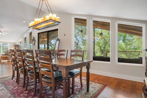 A dining area with a wooden table and chairs, large windows, and a ceiling light fixture. - Rockin B Creek View | 3BR Stay w/ Creek & Kayaks (Pipe Creek)