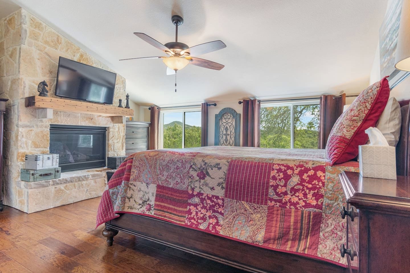 A bedroom with a stone fireplace, a large bed with a patchwork quilt, a ceiling fan, and a television mounted on the wall.