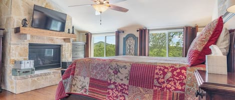A bedroom with a stone fireplace, a large bed with a patchwork quilt, a ceiling fan, and a television mounted on the wall.