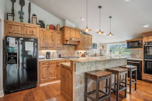 A modern kitchen with wooden cabinets, a stainless steel refrigerator, a stone island with bar stools, and pendant lighting. - Rockin B Creek View | 3BR Stay w/ Creek & Kayaks (Pipe Creek)
