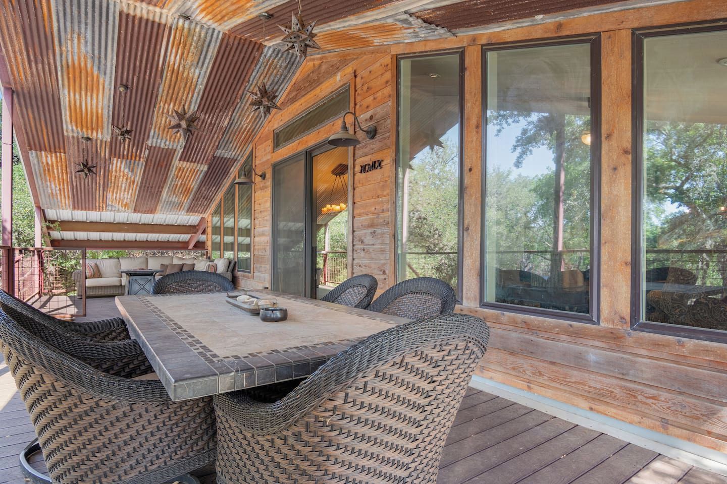 Une terrasse en bois avec une table à manger et des chaises en osier, de grandes fenêtres et un luminaire en forme d’étoile.
