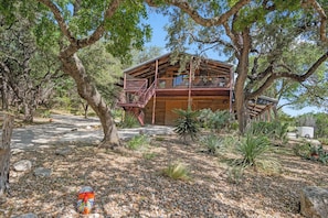 A wooden cabin with a balcony, surrounded by trees and pebbles. - Rockin B Creek View | 3BR Stay w/ Creek & Kayaks (Pipe Creek)