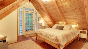 A cozy attic bedroom with a sloped ceiling, a bed, a nightstand, a lamp, and a window with blinds. - Cozy, clean, updated cabin w/hot tub, fireplace, fire pit, BBQ, smart TV (Packwood)