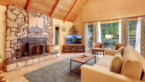 A cozy living room with a stone fireplace, a television, a sofa, and a large window with curtains. - Cozy, clean, updated cabin w/hot tub, fireplace, fire pit, BBQ, smart TV (Packwood)