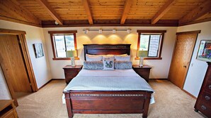 A bedroom with a wooden ceiling, a large bed with multiple pillows, two bedside tables with lamps, and two windows. - Ocean views. Long stay discount. Short walk to Shell beach, dog friendly. (Sea Ranch)