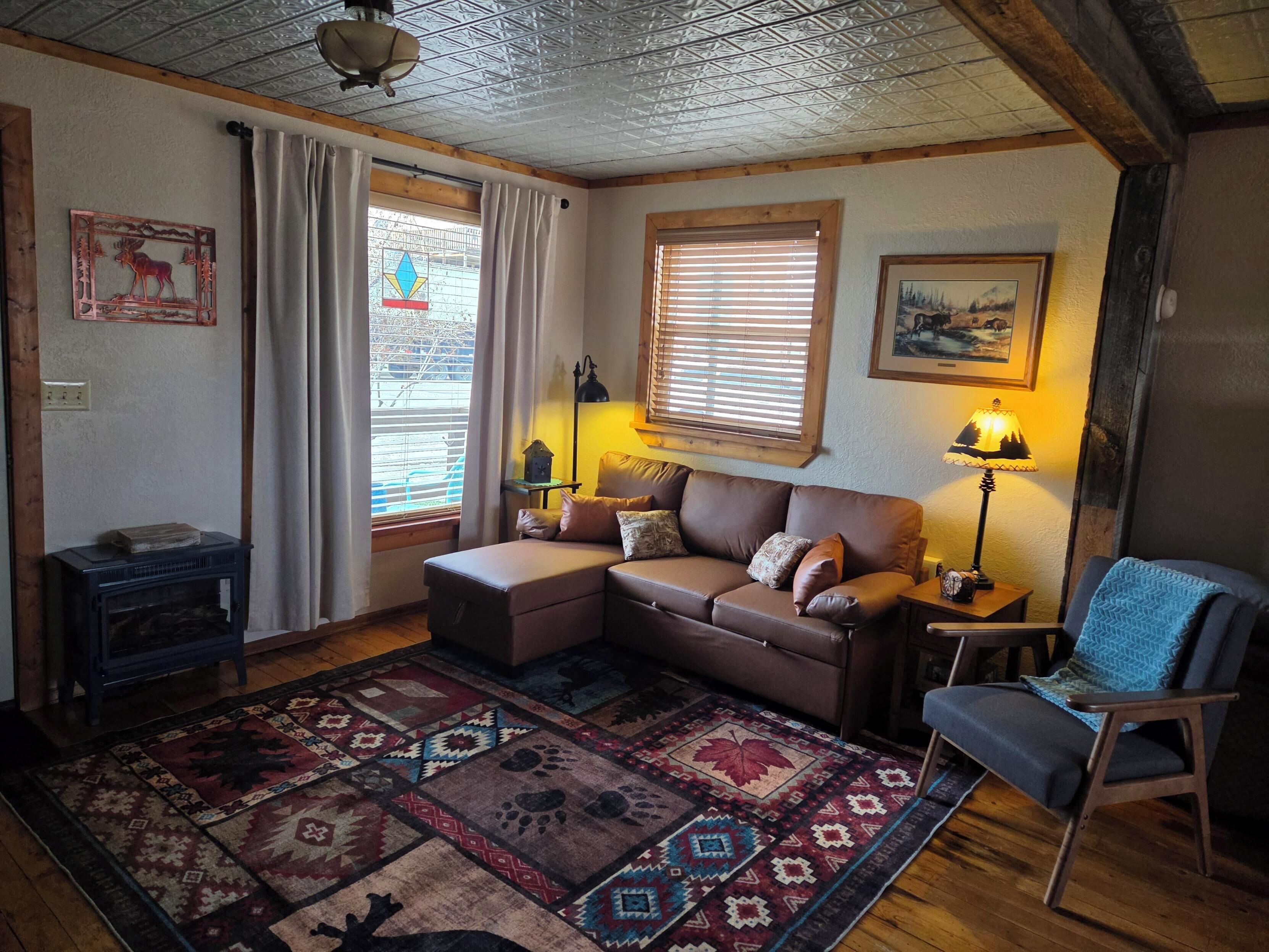 Un salon chaleureux avec un canapé marron, un fauteuil bleu, un tapis à motifs, une cheminée et une fenêtre avec des stores.